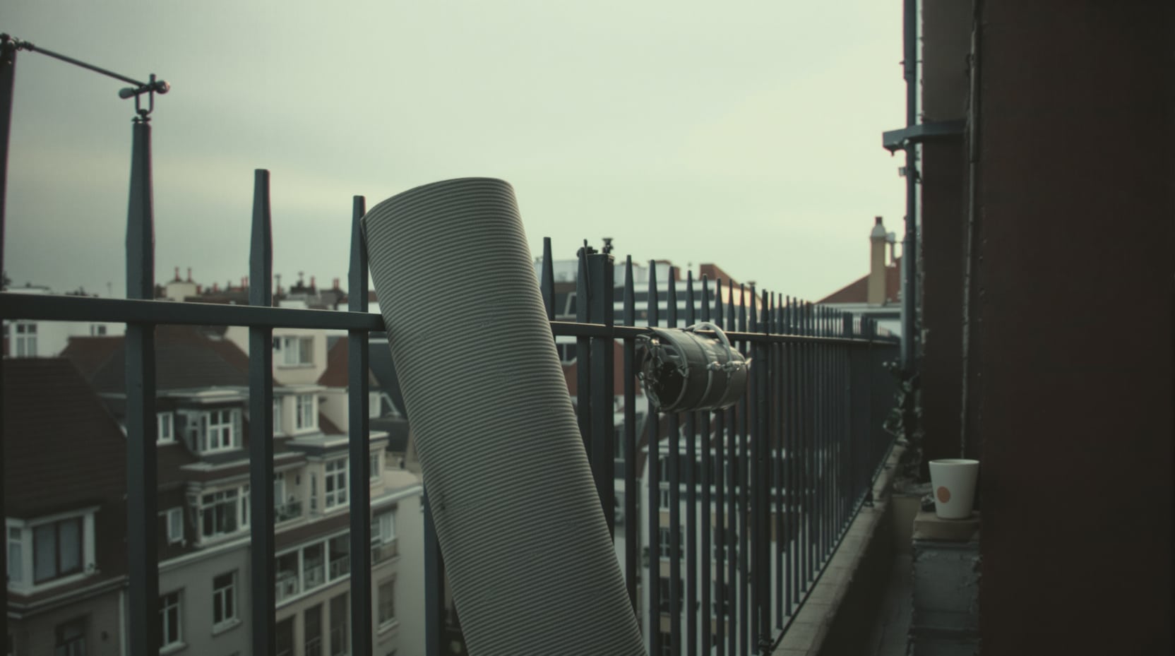 Wide quiet scene of a small city apartment balcony at early dawn, a rolled grey exercise mat leaning on a metal railing,